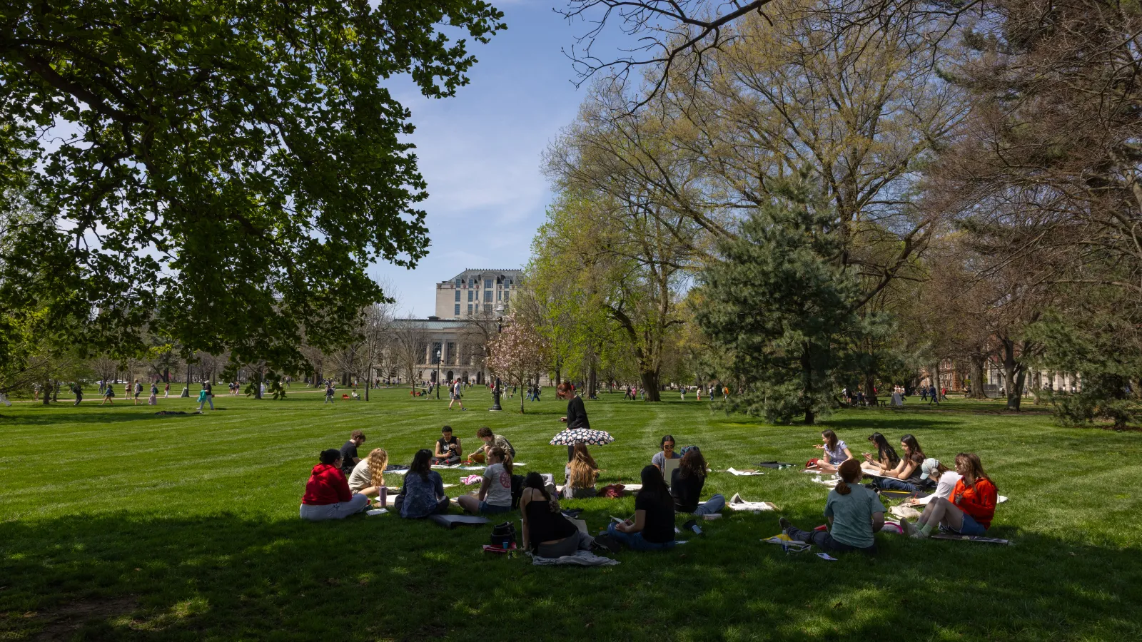People sitting on the oval in the sun in front of Thompson Library
