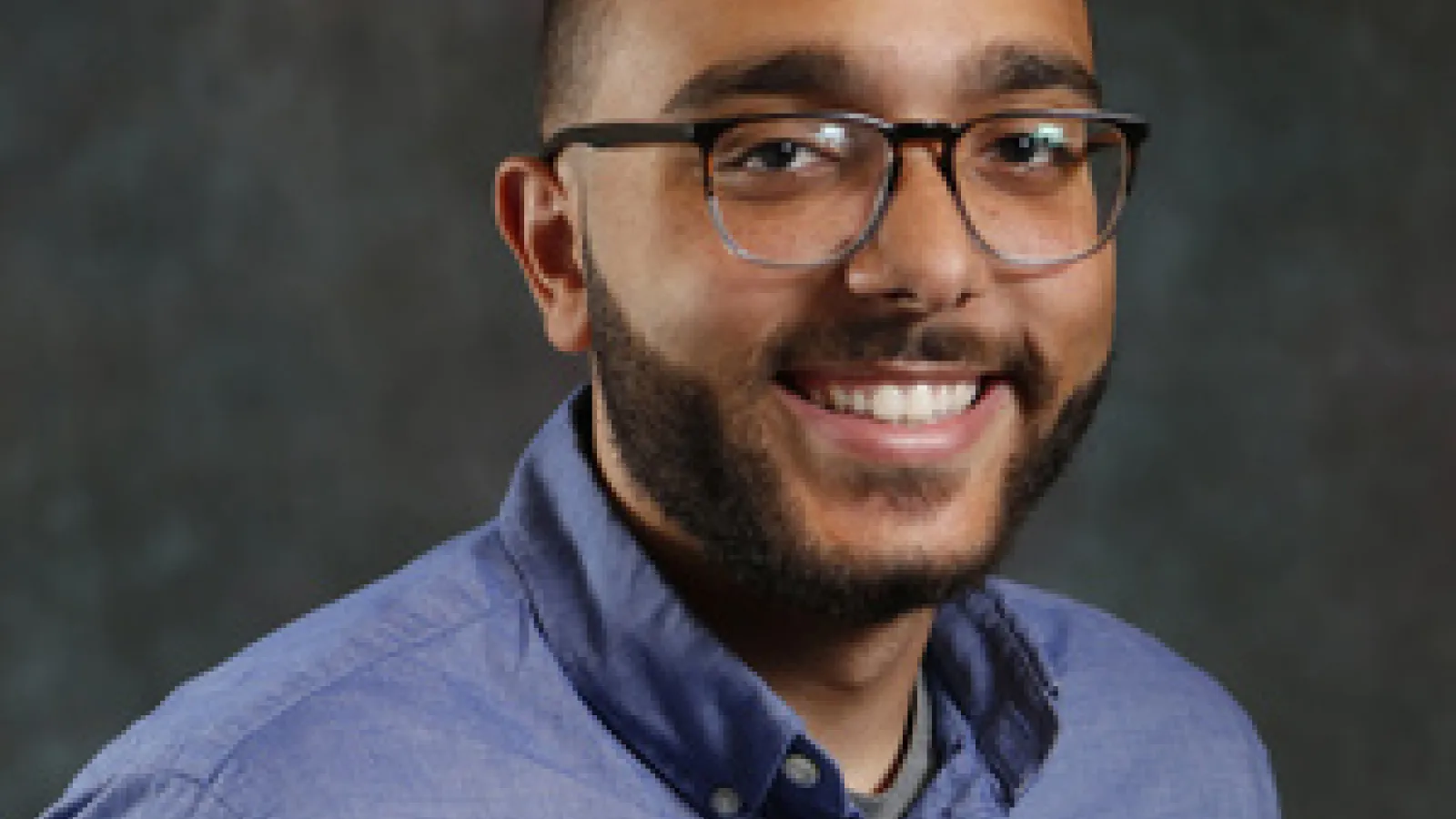 Black male smiling at camera, wearing a blue shirt and glasses
