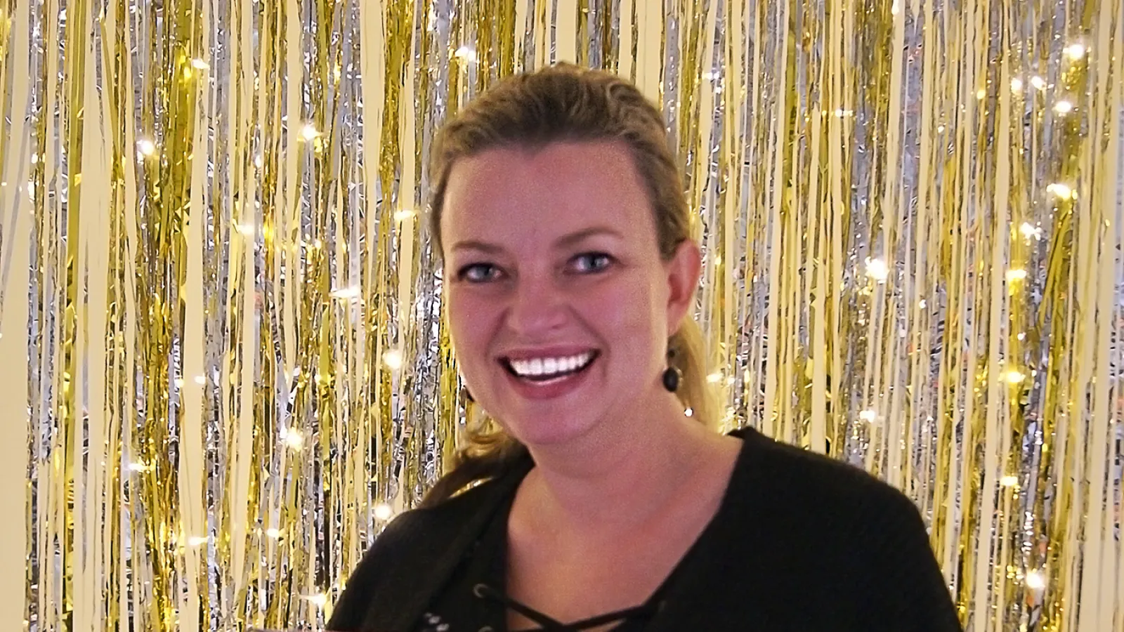 White female, holding glass award, standing in from of strung lights and tinsel.