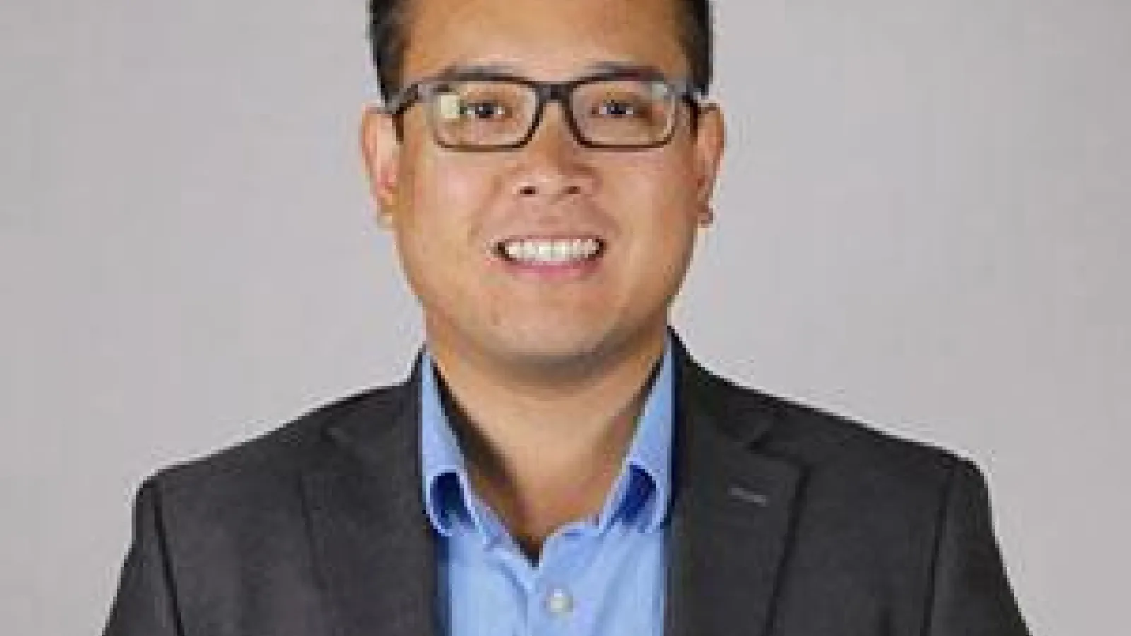 Image of an Asian man with dark short hair and glasses, wearing a black suit with a blue tie smiling at the camera.