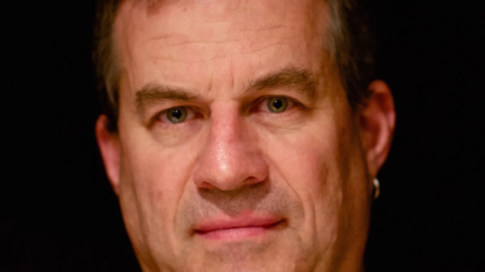 Image of a white man, looking at camera with a flat look on his face, against a back background