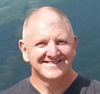 Older white man, wearing a black shirt, standing in front of a green landscape, smiling at camera