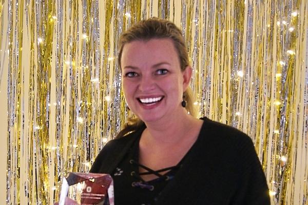 White female, holding glass award, standing in from of strung lights and tinsel.