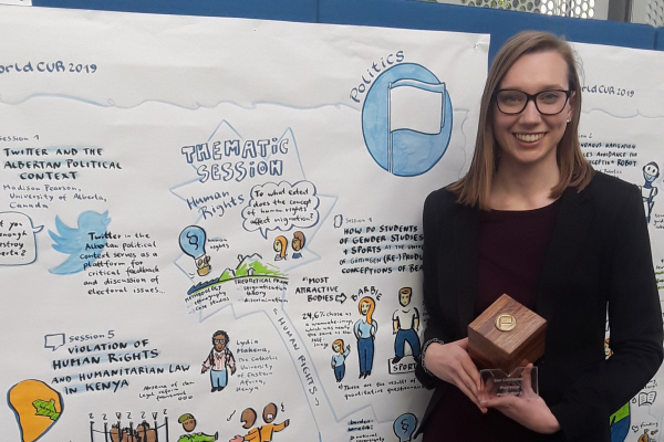 Blond woman wearing black jacket standing in front of presentation board holding small award