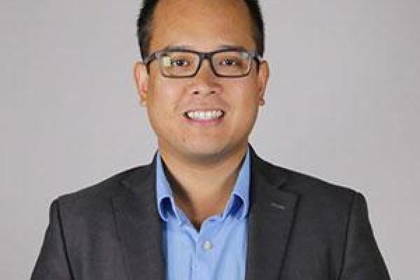 Image of an Asian man with dark short hair and glasses, wearing a black suit with a blue tie smiling at the camera.